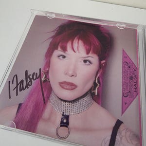 Puede incluir: Una funda de CD con un retrato de Halsey con cabello rosado y un collar de plata. La portada del álbum incluye la firma del artista y el texto "THE GREAT IMERSONATOR HALSEY". El fondo es de color rosa claro.