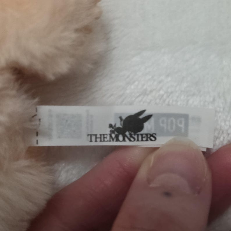 Op de afbeelding: Een wit stoffen label bevestigd aan een beige pluche speelgoed. Het label bevat de zwarte tekst THE MONSTERS, een kleine zwarte afbeelding van een wezen met lange oren en de gedeeltelijke tekst 909 op een lichtgrijze achtergrond rechts.