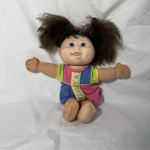 May include: A vintage Cabbage Patch Kids doll with brown hair styled in two high pigtails. The doll has blue eyes and is wearing a colorful outfit with pink, blue, yellow, and green accents. The doll is sitting with its arms outstretched.