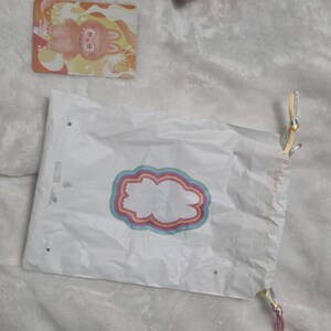 Op de afbeelding: Een witte plastic tas met trekkoord voorzien van een centrale afbeelding van een wit silhouet omringd door concentrische regenboogkleurige ringen. Naast de tas ligt een rechthoekige kaart met een cartoonfiguur met oranje vacht.