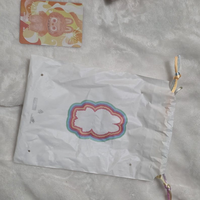 Op de afbeelding: Een witte plastic tas met trekkoord voorzien van een centrale afbeelding van een wit silhouet omringd door concentrische regenboogkleurige ringen. Naast de tas ligt een rechthoekige kaart met een cartoonfiguur met oranje vacht.