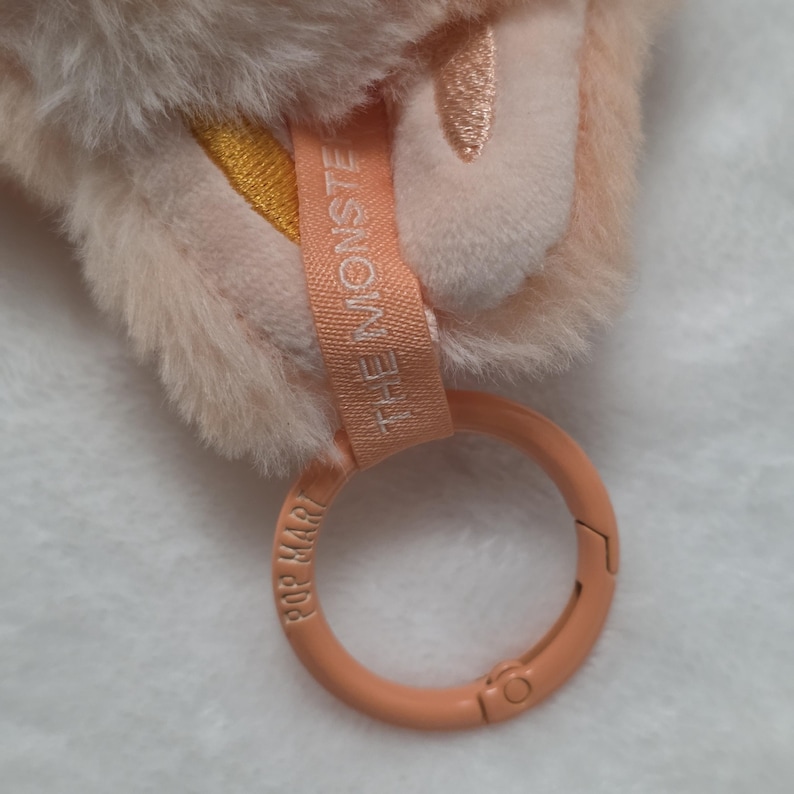 Op de afbeelding: Een perzikkleurige pluche sleutelhanger met zachte vacht, een stoffen label bedrukt met de tekst THE MONSTERS en een ronde perzikkleurige plastic ring met de tekst POP MART in reli&euml;f. Het ontwerp bevat geborduurde gele en roze accenten.