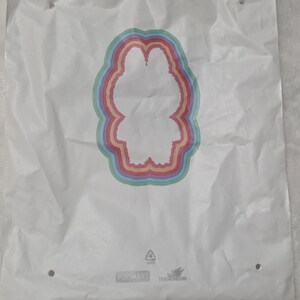Op de afbeelding: Een witte plastic tas met een centraal silhouet van een personage omringd door concentrische regenboogkleurige ringen. Onderaan de tas staat de tekst POP MART en THE MONSTERS samen met een recyclingsymbool.