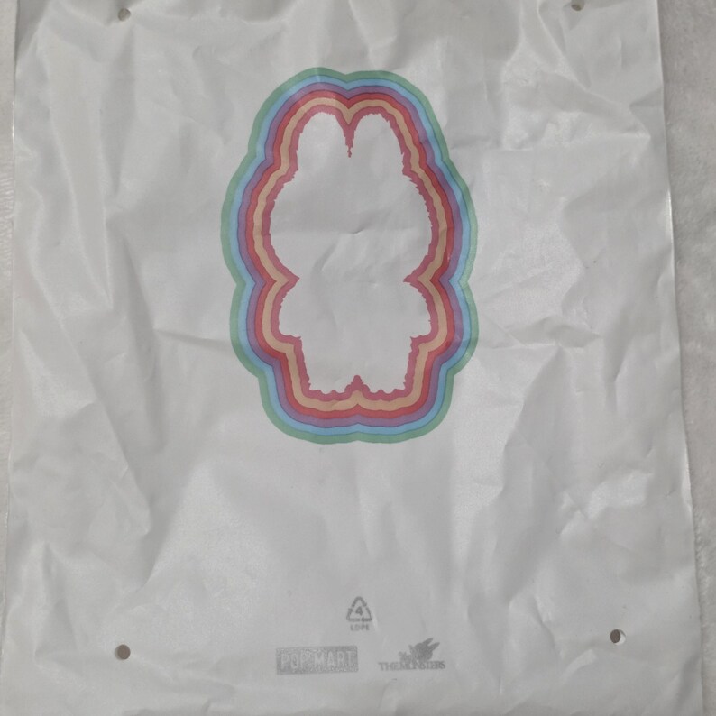 Op de afbeelding: Een witte plastic tas met een centraal silhouet van een personage omringd door concentrische regenboogkleurige ringen. Onderaan de tas staat de tekst POP MART en THE MONSTERS samen met een recyclingsymbool.