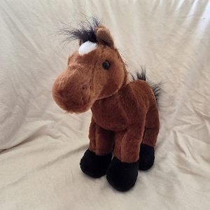 Op de afbeelding: Een bruin pluche paardenspeelgoed met een witte vlek op het voorhoofd en zwarte accenten op de manen, staart en hoeven. Het knuffeldier staat rechtop en heeft een vriendelijke, cartoonachtige uitstraling.