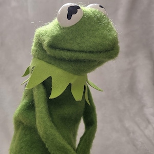 May include: A green felt frog puppet featuring large white eyes with black pupils, a wide mouth, and a jagged light green collar around the neck. The puppet has a textured, fuzzy surface and is positioned in a profile view against a neutral background.