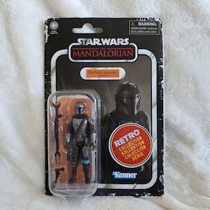 May include: A Star Wars The Mandalorian action figure in a retro collection package. The figure is silver and black, with a helmet and armor. The packaging includes the text "The Mandalorian (Beskar)" and "Retro Collection".