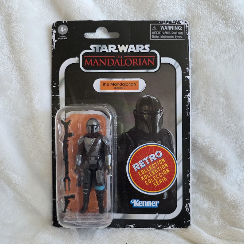 May include: A Star Wars The Mandalorian action figure in a retro collection package. The figure is silver and black, with a helmet and armor. The packaging includes the text "The Mandalorian (Beskar)" and "Retro Collection".