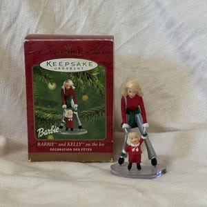 Peut inclure: Ornement de Noël Barbie et Kelly sur la glace. L'ornement représente deux figurines en tenues rouges et vertes sur des patins à glace. La boîte est rouge avec des lettres dorées et les mots "Keepsake Ornament" et "Barbie".