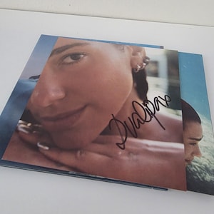 Pode incluir: Uma capa de álbum assinada com um retrato em close-up. A assinatura, "Dua Lipa", está em tinta preta. A capa do álbum tem um esquema de cores azul e bege.
