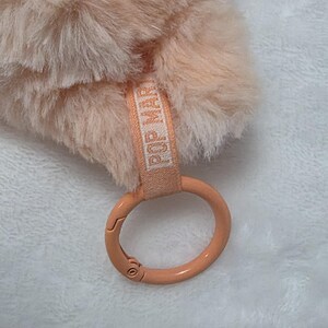 Op de afbeelding: Een perzikkleurig accessoire van imitatiebont met een zachte, pluizige textuur. Bevestigd is een licht perzikkleurige stoffen band met de tekst POP MART in het wit gedrukt, verbonden aan een gladde, ronde, perzikkleurige metalen karabijnring.