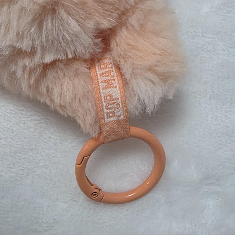 Op de afbeelding: Een perzikkleurig accessoire van imitatiebont met een zachte, pluizige textuur. Bevestigd is een licht perzikkleurige stoffen band met de tekst POP MART in het wit gedrukt, verbonden aan een gladde, ronde, perzikkleurige metalen karabijnring.