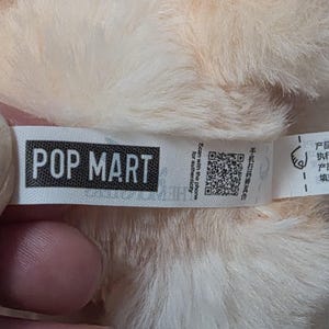 Op de afbeelding: Een wit stoffen label bevestigd aan zacht, licht perzikkleurig pluche materiaal. Het label bevat de zwarte tekst POP MART, een QR-code voor authenticiteit en de Engelse zin Scan with the phone for authenticity naast Chinese karakters.