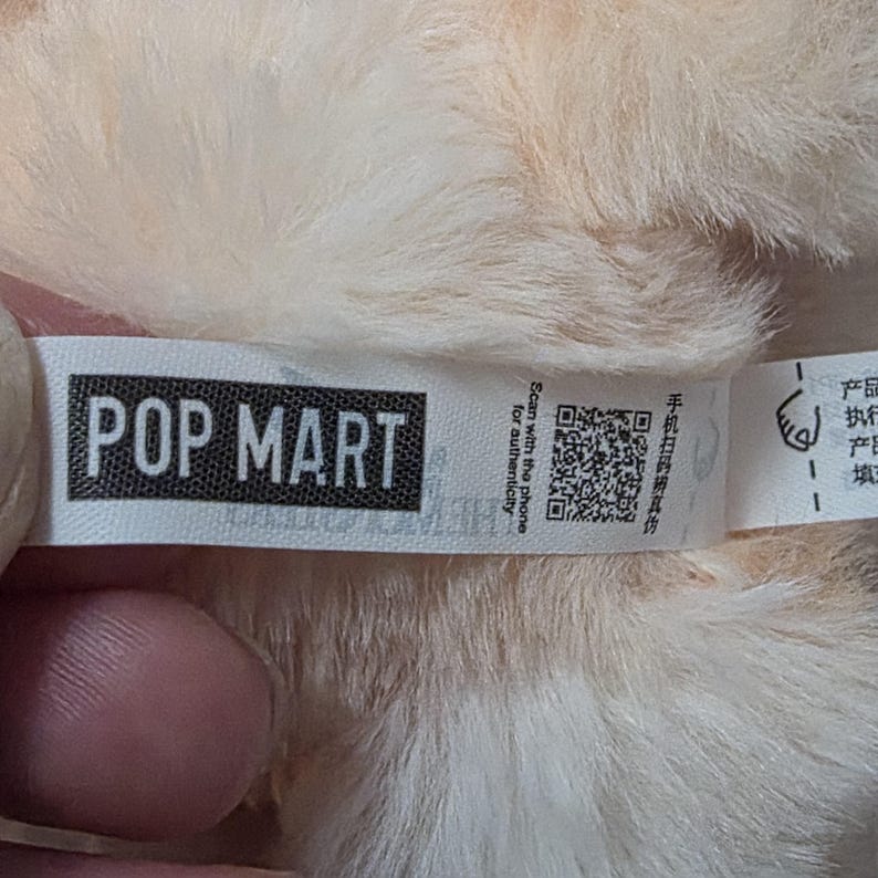 Op de afbeelding: Een wit stoffen label bevestigd aan zacht, licht perzikkleurig pluche materiaal. Het label bevat de zwarte tekst POP MART, een QR-code voor authenticiteit en de Engelse zin Scan with the phone for authenticity naast Chinese karakters.
