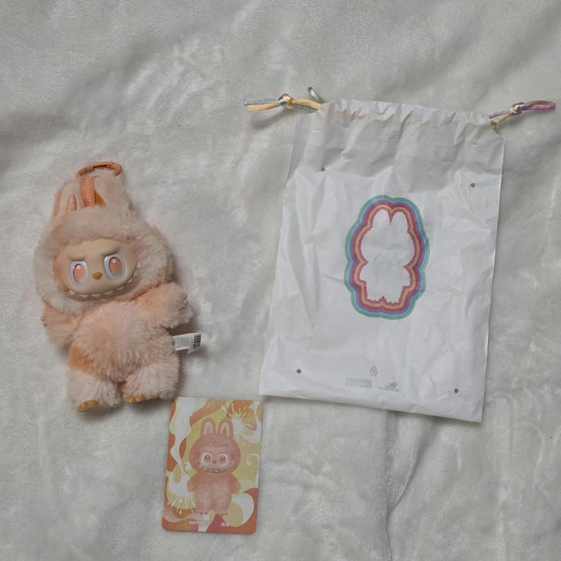 Op de afbeelding: Een perzikkleurig pluche wezen met een capuchon, grote oranje ogen en een grijns met tanden. Daarnaast ligt een witte tas met trekkoord met een kleurrijke, gelaagde omtrek van het wezen en een rechthoekige kaart met een illustratie van het figuur.