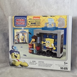 Könnte beinhalten: Ein Mega Bloks The SpongeBob Movie Sponge Out of Water Photo Booth Time Machine Baukasten. Die Verpackung zeigt eine gelbe SpongeBob-Figur, eine baubare graue Kabine, ein Kelp Soda-Fass und eine Uhr. Das Set enthält insgesamt 98 Teile.