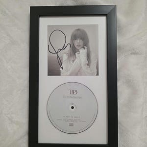 Pode incluir: Uma moldura retangular preta contendo uma fotografia a preto e branco de Taylor Swift com uma assinatura a tinta preta sobre um CD circular. O CD apresenta o texto TTPD, The Tortured Poets Department e Taylor Swift impresso a preto.