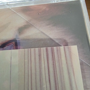 Pu&ograve; includere: Una custodia per CD sigillata con una superficie argentata riflettente. L'involucro di plastica &egrave; visibile, con un angolo di un foglio a strisce bianche e rosse che spunta da sotto. L'immagine &egrave; leggermente sfocata.
