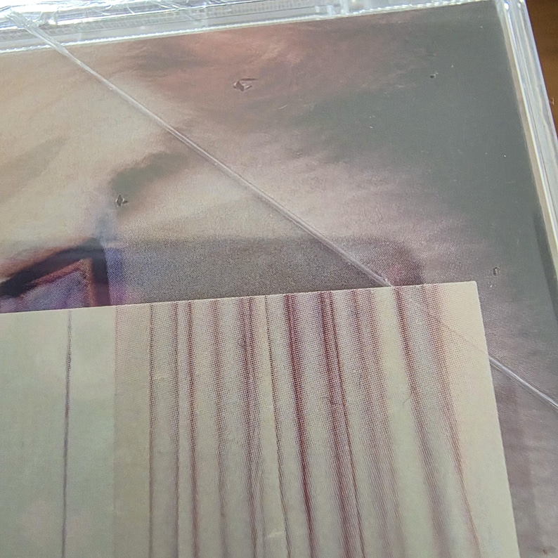 Pu&ograve; includere: Una custodia per CD sigillata con una superficie argentata riflettente. L'involucro di plastica &egrave; visibile, con un angolo di un foglio a strisce bianche e rosse che spunta da sotto. L'immagine &egrave; leggermente sfocata.