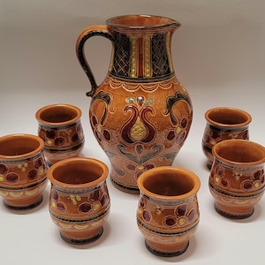 Pode incluir: Um jarro de cerâmica e seis xícaras combinando. O conjunto apresenta uma cor de base marrom com desenhos florais e geométricos em vermelho, amarelo e preto. O jarro tem uma alça grande e um topo decorativo.