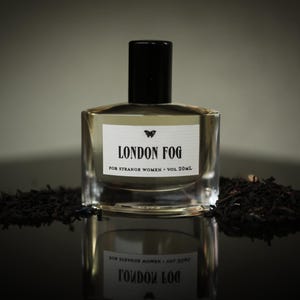 May include: A perfume bottle with a black cap and a label reading "LONDON FOG" with a butterfly graphic. The bottle is on a reflective surface, with dark tea leaves around it. The label says "FOR STRANGE WOMEN - VOL 20ml."