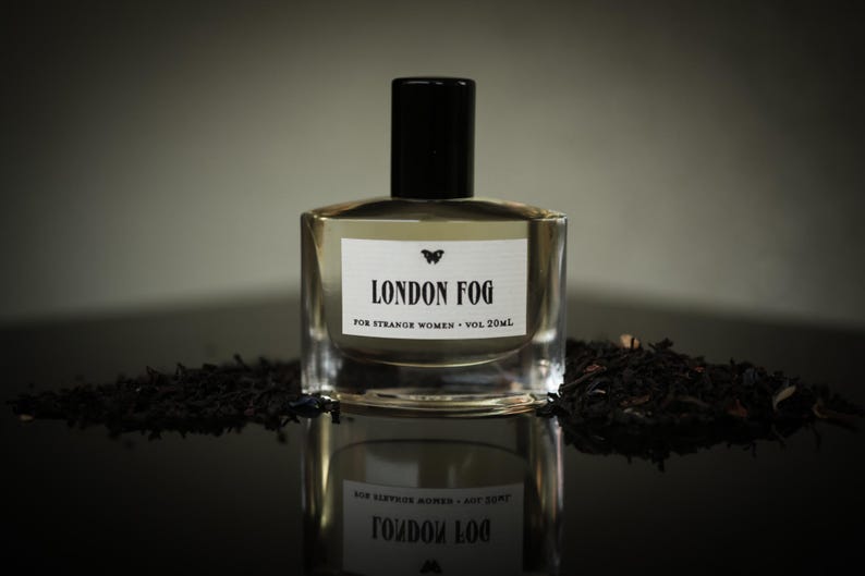 May include: A clear glass perfume bottle with a black cap, labeled "LONDON FOG" with a butterfly graphic. The bottle sits on a reflective surface, surrounded by dark tea leaves. The label reads "FOR STRANGE WOMEN - VOL 20ml."