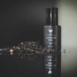 May include: A black bottle of "London Fog" tea with a black cap, and a pile of loose tea leaves. The bottle has white text and a butterfly logo. The bottle and tea are reflected on a dark surface.