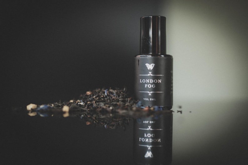 May include: A black bottle of "London Fog" tea with a black cap, and a pile of loose tea leaves. The bottle has white text and a butterfly logo. The bottle and tea are reflected on a dark surface.
