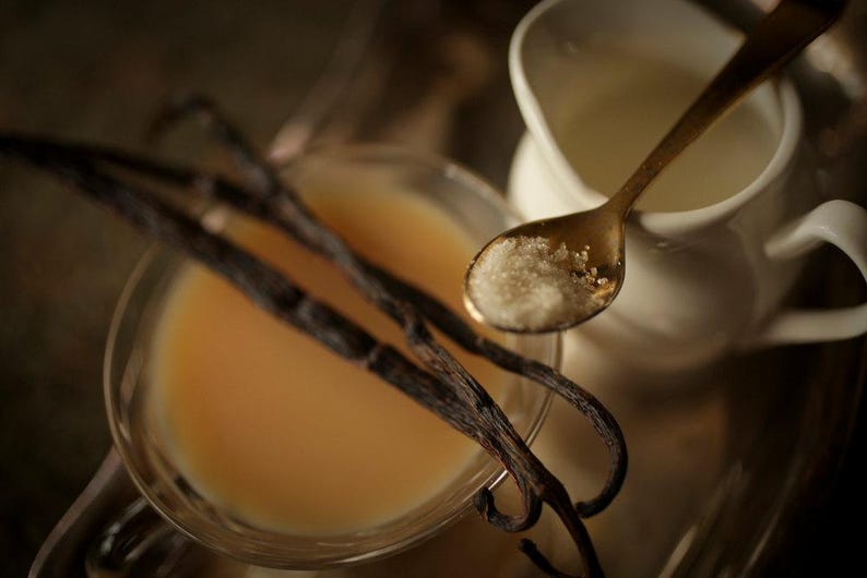 May include: A still life image featuring a clear glass bowl with a light orange liquid, likely a custard or sauce, and several dark brown vanilla beans. A small white creamer pitcher and a gold spoon with sugar complete the composition.