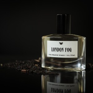 May include: A clear glass perfume bottle with a black cap, labeled "LONDON FOG" with a butterfly logo. The bottle sits on a dark surface with a pile of dark tea leaves. The text reads "FOR STRANGE WOMEN &bull; VOL 20ml."