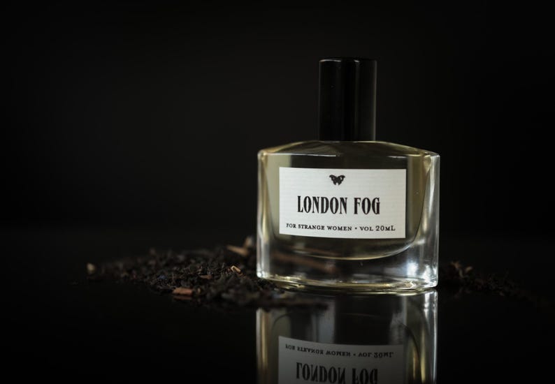 May include: A clear glass perfume bottle with a black cap, labeled "LONDON FOG" with a butterfly logo. The bottle sits on a dark surface with a pile of dark tea leaves. The text reads "FOR STRANGE WOMEN &bull; VOL 20ml."
