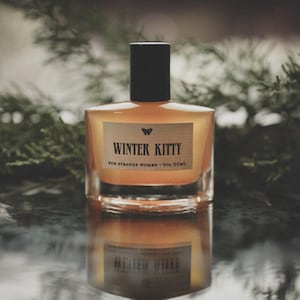 May include: A square glass perfume bottle with a black cap and a light orange liquid inside. The label reads "WINTER KITTY" with a butterfly graphic. The bottle is reflected on a dark, glossy surface, with greenery in the background.