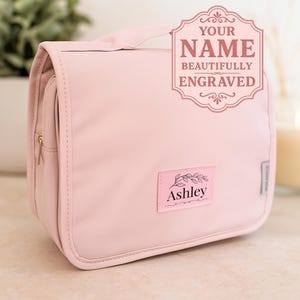May include: A light pink hanging toiletry bag with a top handle and a gold-tone zipper. A rectangular pink patch on the front features a black leaf graphic and the name Ashley. A decorative graphic overlay in the corner reads YOUR NAME BEAUTIFULLY ENGRAVED.