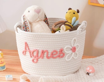 Könnte beinhalten: Ein weißer Seilkorb mit dem Namen "Agnes" in rosa Schrift und einer Blumenapplikation. Der Korb enthält ein Stoffkaninchen mit floralen Ohren und ein gelb-braunes Bienenspielzeug. Der Korb hat zwei Griffe.