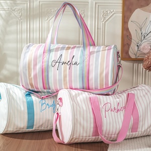 Puede incluir: Tres bolsas de lona a rayas en colores pastel, cada una con un nombre bordado en la parte delantera. La bolsa superior tiene una correa arcoíris y el nombre "Amelia". Las otras bolsas tienen correas azules y rosas, con los nombres "Bridg" y "Parker".