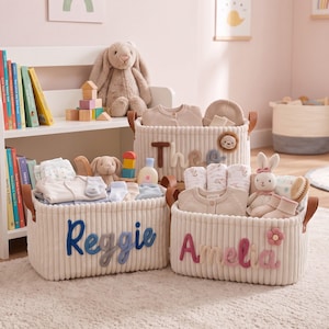 Puede incluir: Tres cestas de almacenamiento de tela acanalada color crema con asas de piel sintética marrón. Las cestas presentan nombres personalizados en letras de hilo en relieve: Reggie en azul, Amelia en rosa con una flor y Theo con una cara de león.
