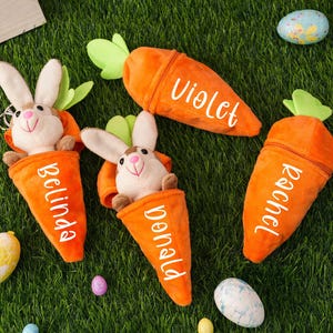 May include: Four plush carrot-shaped Easter baskets with bunny faces and personalized names: Belinda, Donald, Rachel, and Violet. The baskets are on a green grassy surface with Easter eggs.