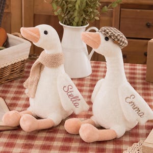 Può includere: Due oche di peluche bianche con becchi e zampe arancioni. Un'oca indossa una sciarpa in maglia beige e ha il nome Stella ricamato sull'ala. L'altra oca indossa un berretto a quadri marrone e bianco con il nome Emma ricamato sull'ala.