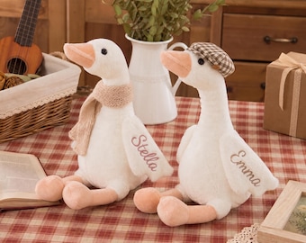 May include: Two plush white goose toys with orange beaks and feet. One goose wears a beige knit scarf and has the name Stella embroidered on its wing. The other goose wears a brown and white plaid cap and has the name Emma embroidered on its wing.
