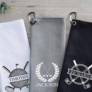 May include: Three golf towels with personalized names and golf-themed designs. The towels are white, gray, and black with a carabiner clip.