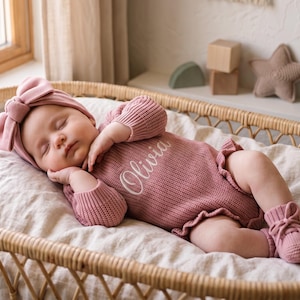 May include: A dusty rose knitted long-sleeve romper with a ruffled hem and the name Olivia embroidered in white script on the chest. The outfit includes a matching dusty rose knitted headband with a large bow and a pair of matching knitted booties.