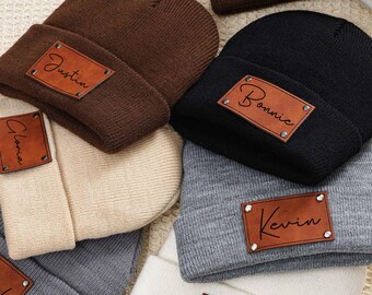 Könnte beinhalten: Eine Sammlung von sechs braunen, schwarzen, beigen und grauen Strickmützen mit Lederpatches mit den Namen Justin, Bonnie, Gloria, Kevin und Hyla.