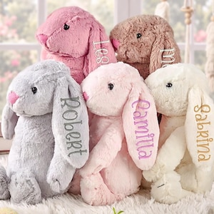 May include: A collection of five plush bunny stuffed animals with soft fur in shades of gray, pink, brown, and cream. Each bunny features long floppy ears, with names embroidered in various thread colors on the ears of the front three bunnies.
