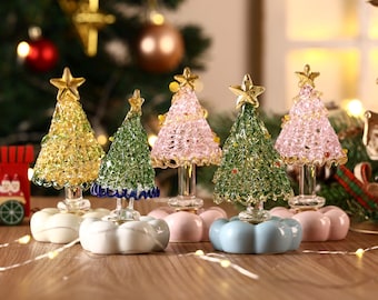 May include: A collection of decorative Christmas trees in various colors, including gold, green, and pink. Each tree is topped with a gold star and sits on a ceramic base. The trees are arranged on a wooden surface with a blurred Christmas tree background.