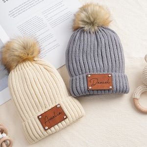 May include: Two knitted baby hats with faux fur pom poms. The hats are gray and cream and have personalized leather name tags that say "Daniel" and "Amelia".