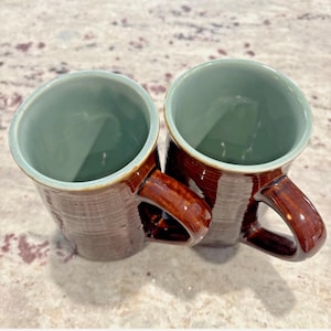 May include: Two ceramic mugs with brown exteriors and handles, and a light green interior. The mugs have a textured surface and are set on a light-colored surface. The handles are a darker shade of brown.
