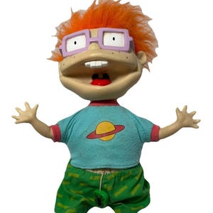 May include: A Chuckie Finster doll with bright orange hair, light purple glasses, and a wide open mouth showing two front teeth. The doll wears a light blue t-shirt with a yellow planet graphic and green pants with a small black spider charm.