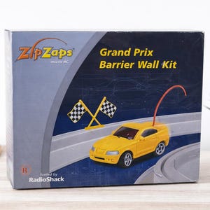 May include: A boxed Zip Zaps Grand Prix Barrier Wall Kit. The box features a yellow toy car, checkered flags, and a race track design. The box is gray and navy blue with the RadioShack logo.