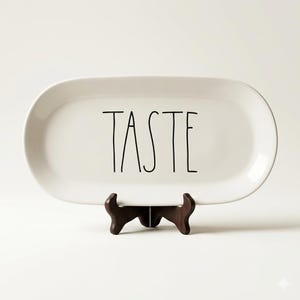 May include: A white, oval-shaped ceramic serving platter with the word "TASTE" in black lettering. The platter is displayed on a dark brown wooden stand, suitable for serving food or as a decorative piece.