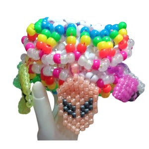Könnte beinhalten: Ein farbenfrohes Perlenarmband mit einem rosafarbenen Alien-Anhänger. Das Armband besteht aus kleinen Plastikperlen in verschiedenen Farben, darunter rosa, lila, blau, grün, gelb und weiß. Der Alien-Anhänger ist eine kleine, rosafarbene Perle mit schwarzen Augen und einem schwarzen Mund.